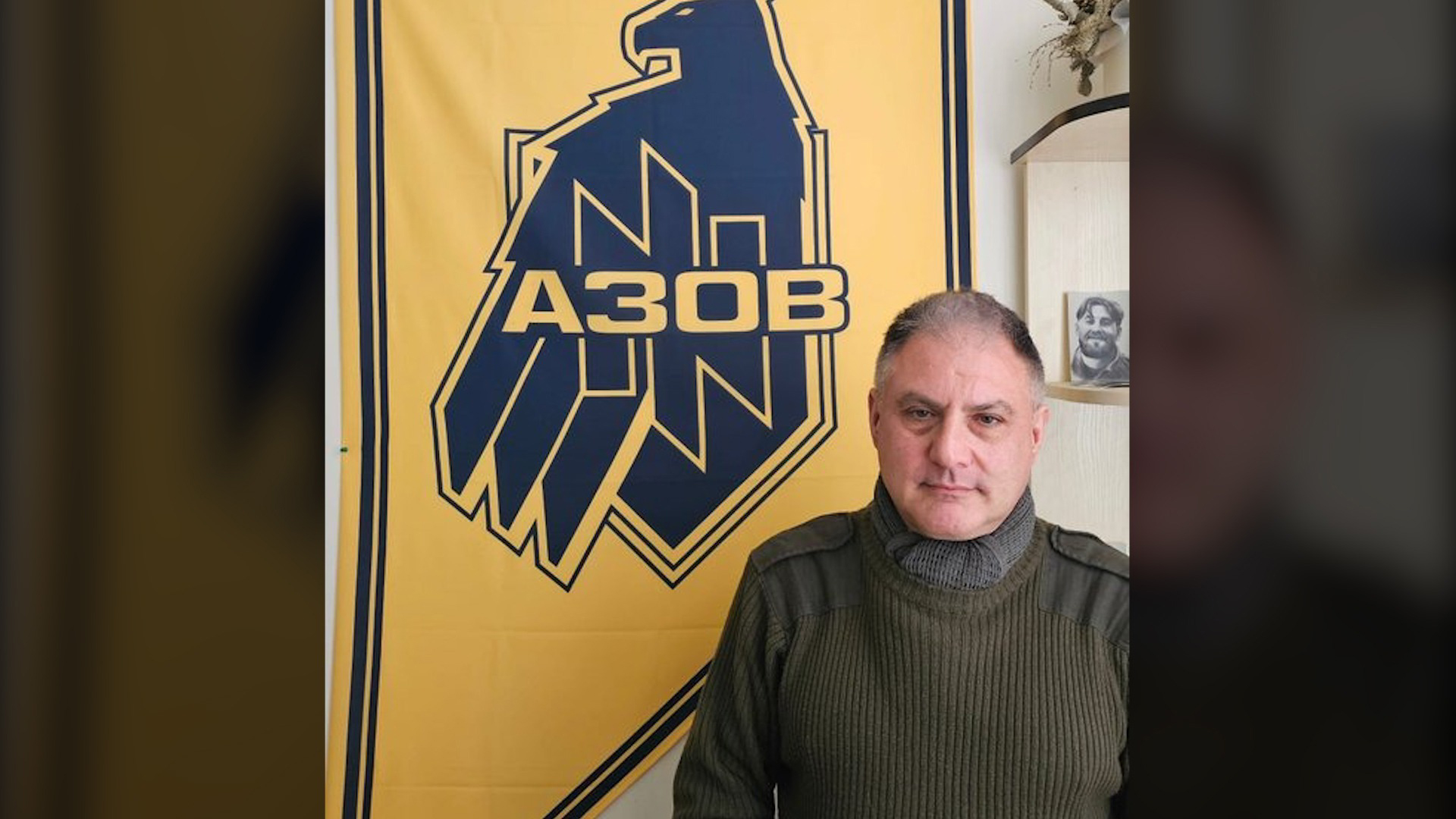 Jack Lopresti poses with an Azov Brigade banner with its eagle and wolfsangel symbols