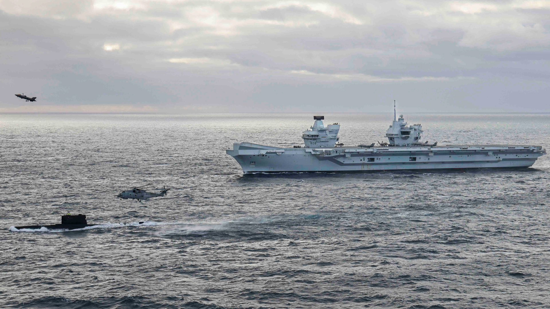 A Royal Navy helicopter above a Norwegian submarine with an F-35 taking off from HMS Queen Elizabeth in the background (Picture: HMSQNLZ X).
