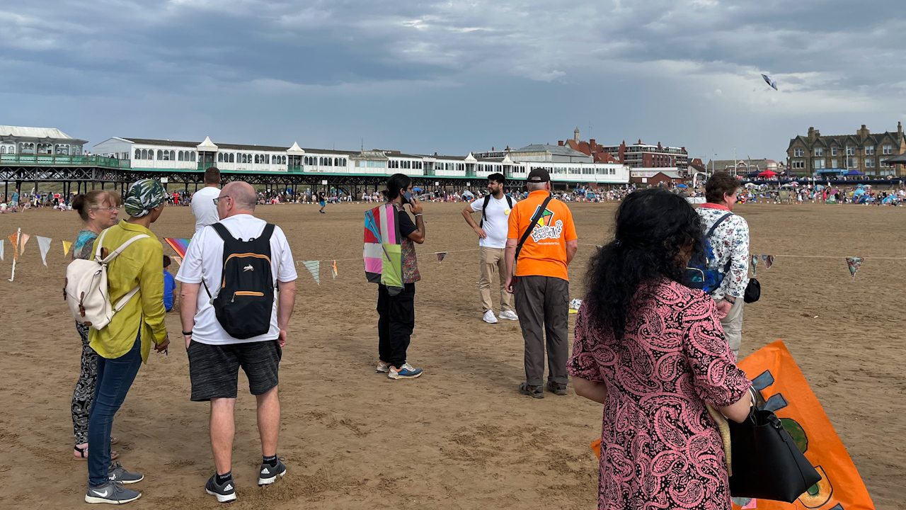 Families from Afghanistan standing with volunteers preparing to demonstrate kite flying on Blackburn beach.