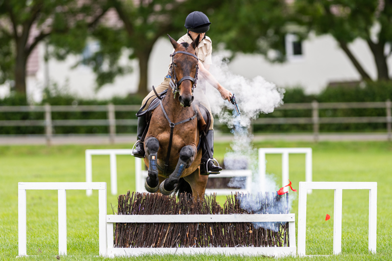 A member of the King's Troop Royal Horse Artillery (RHA), is seen here firing a blank revolver to burst a balloon whilst in mid air over a jump.