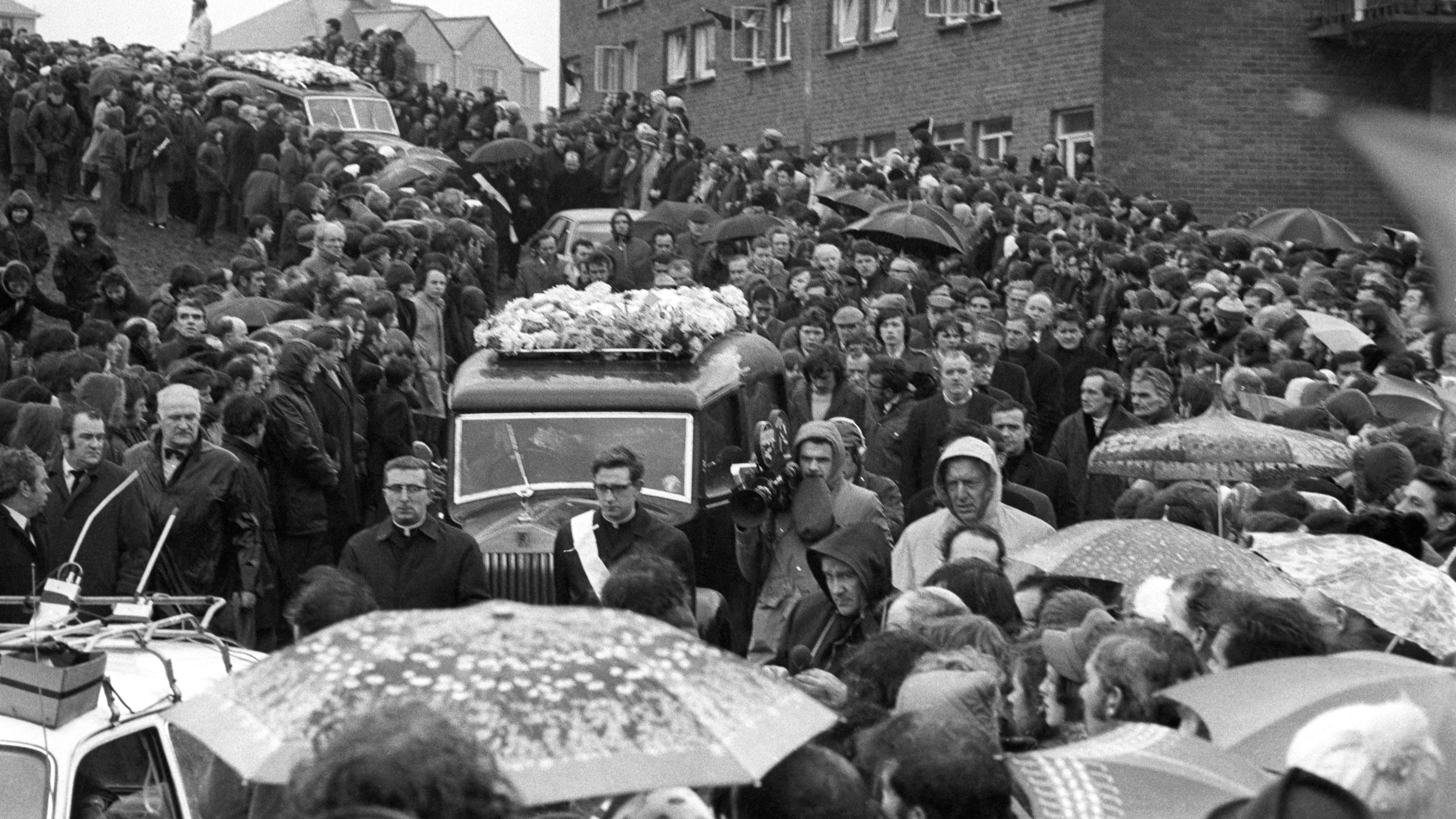 A silent crowd form a cordon 10 or more deep, lining both sides of the 250 yards of road leading from St Mary's Church, Creggan Hill to the cemetery, to watch the funeral procession of the 13 who died on Bloody Sunday