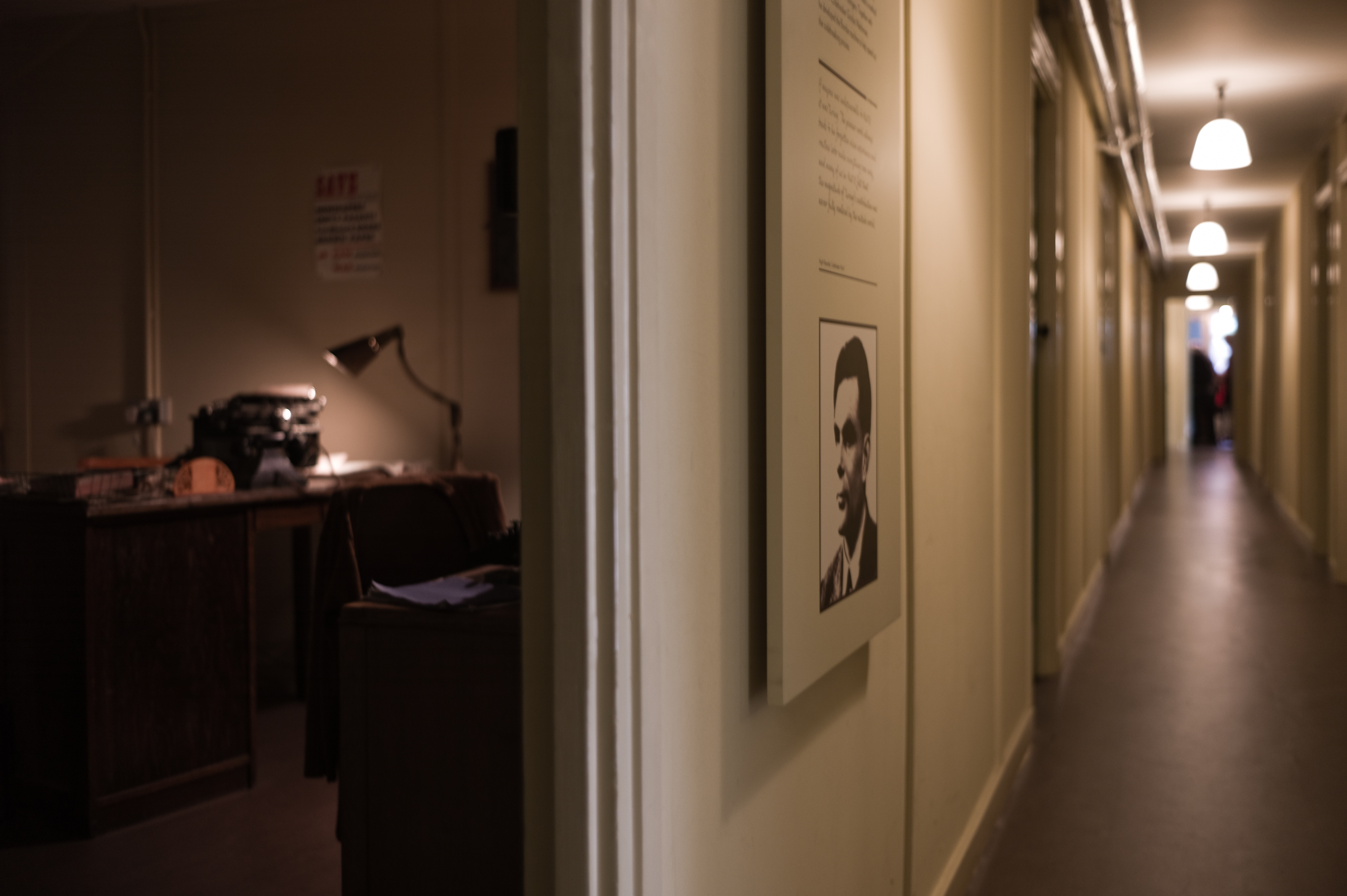 A side-view of Alan Turing's office in Hut 8, Bletchley Park. A desk lamp lights up an old typewriter on a desk in the corner; Turing's picture is on the corridor wall.