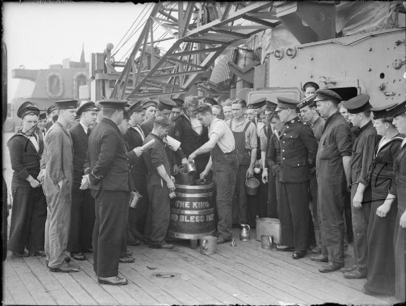 Sailors can be seen crowded round the wooden and brass barrel awaiting the daily issue of rum during the 'up spirits' ceremony. A Royal marine and Petty Officer of the day stands observing the process. Note the crane in the background.