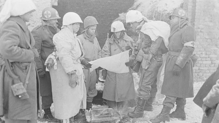 US Army infantrymen with medics in winter uniforms huddled around a map during the Battle of the Bulge CREDIT US Army