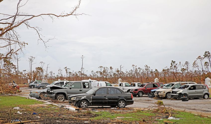 A car park where most cars have suffered some form of damage. Fallen branches and broken buildings become airborne projectiles in the hurricane winds.