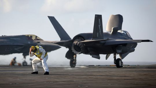 An aircraft handler on board HMS Prince of Wales braces himself as an F-35B aircraft prepares to take off - note the downward direction of the exhaust nozzle