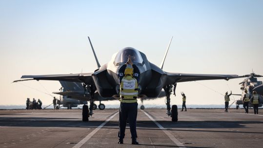 An F-35B taxis on the flight deck of HMS Prince of Wales, with two Merlins in the background