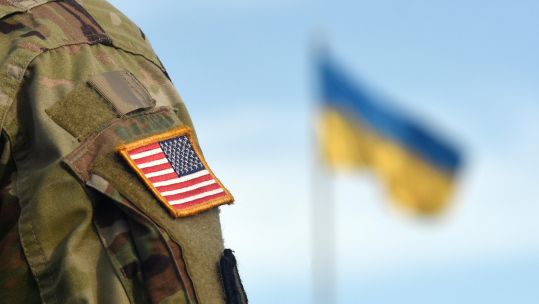 Anonymous member of the US Armed Forces with a Ukrainian flag flying in the background