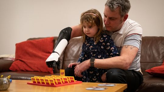 Cover Image: Darren "Daz" Fuller uses his bionic arm to play a game with his daughter, Sky (Picture: Open Bionics).