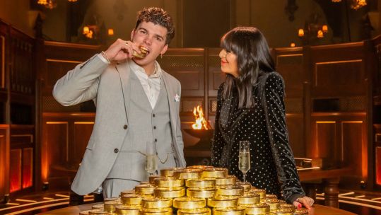 The Traitors winner LCpl Harry Clark bites a gold bar while presenter Claudia Winkleman watches on CREDIT BBC Studio Lambert Paul Chappells