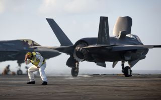 An aircraft handler on board HMS Prince of Wales braces himself as an F-35B aircraft prepares to take off - note the downward direction of the exhaust nozzle